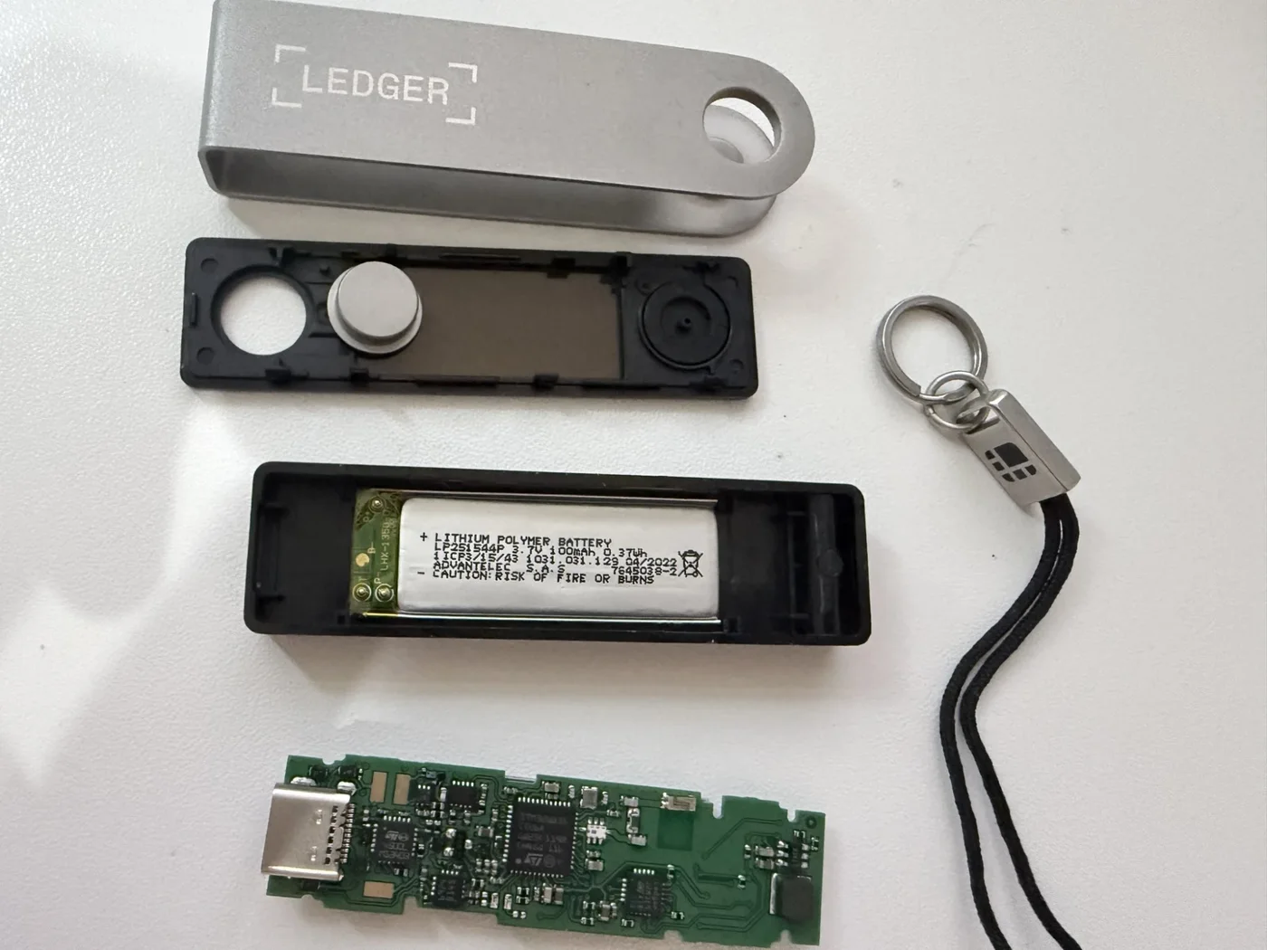 Interior de un Ledger Nano X intacto, cara de componentes: una placa de circuito verde con el chip principal, un puerto USB-C y chips pequeños, junto a la carcasa negra abierta con su batería de polímero de litio, la cubierta metálica y el llavero
