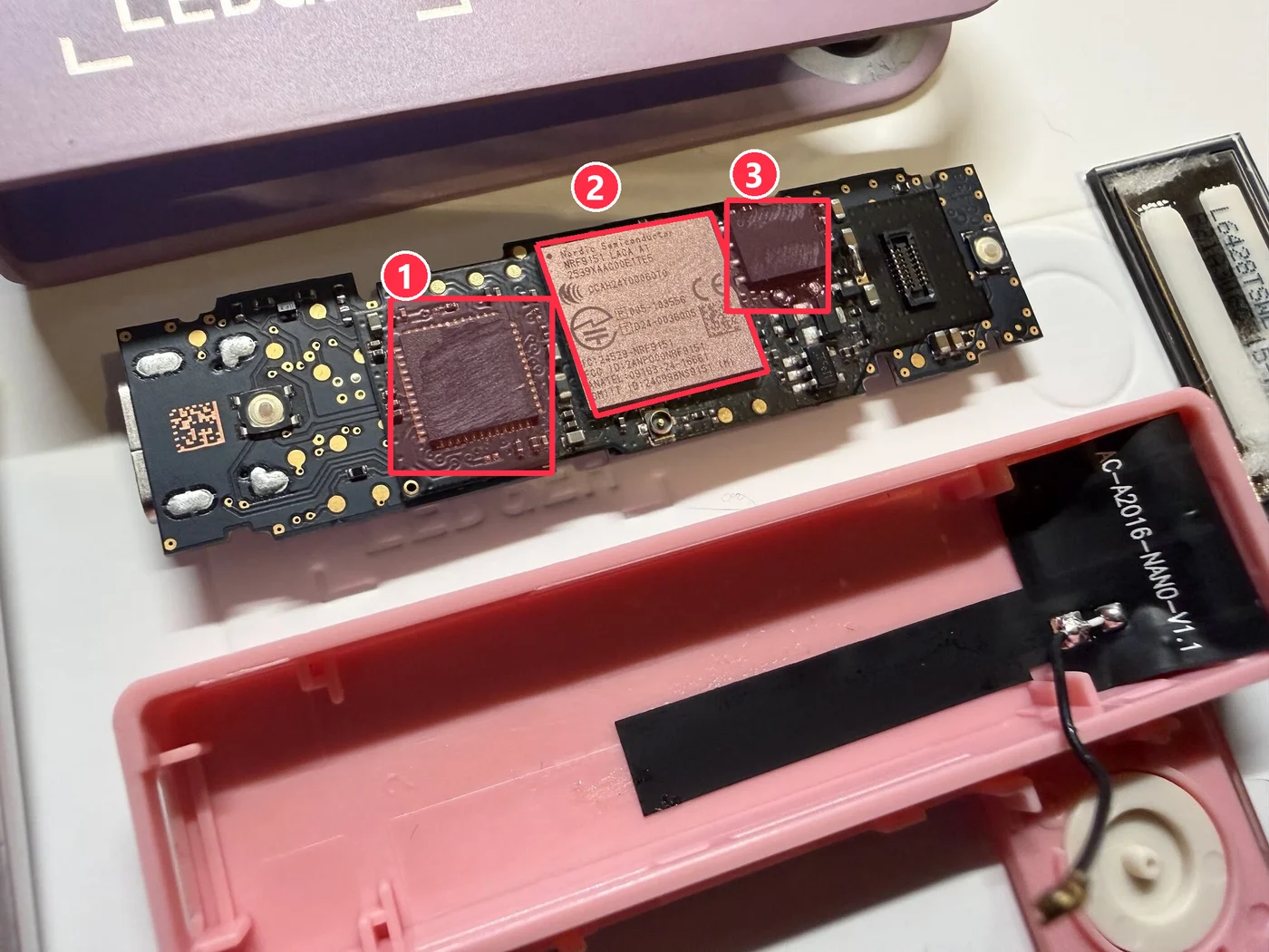 Foto de la cara de la pantalla de un Ledger Nano X manipulado con la pantalla levantada, con tres piezas rodeadas en rojo: 1 un chip a la izquierda con las marcas lijadas, 2 un módulo celular Nordic nRF9151 en el centro, 3 un segundo chip a la derecha junto al conector de la pantalla